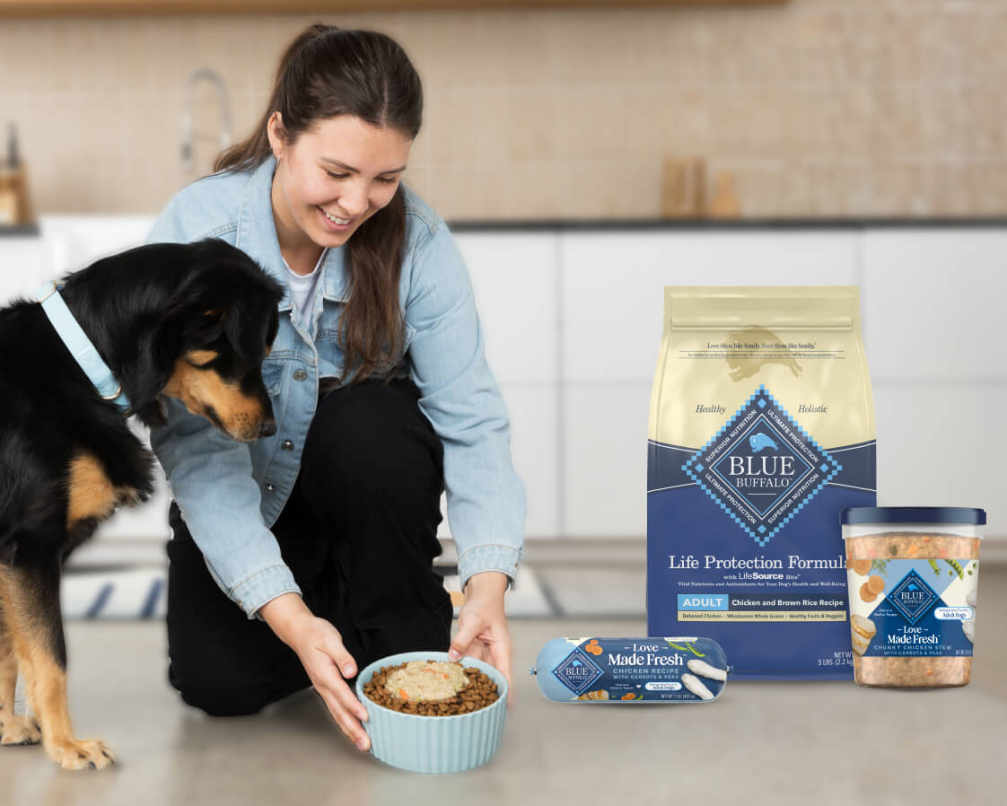 Woman kneels to set down a dog food bowl as a dog watches; Blue Buffalo Life Protection Formula bag with Love Made Fresh roll and tub