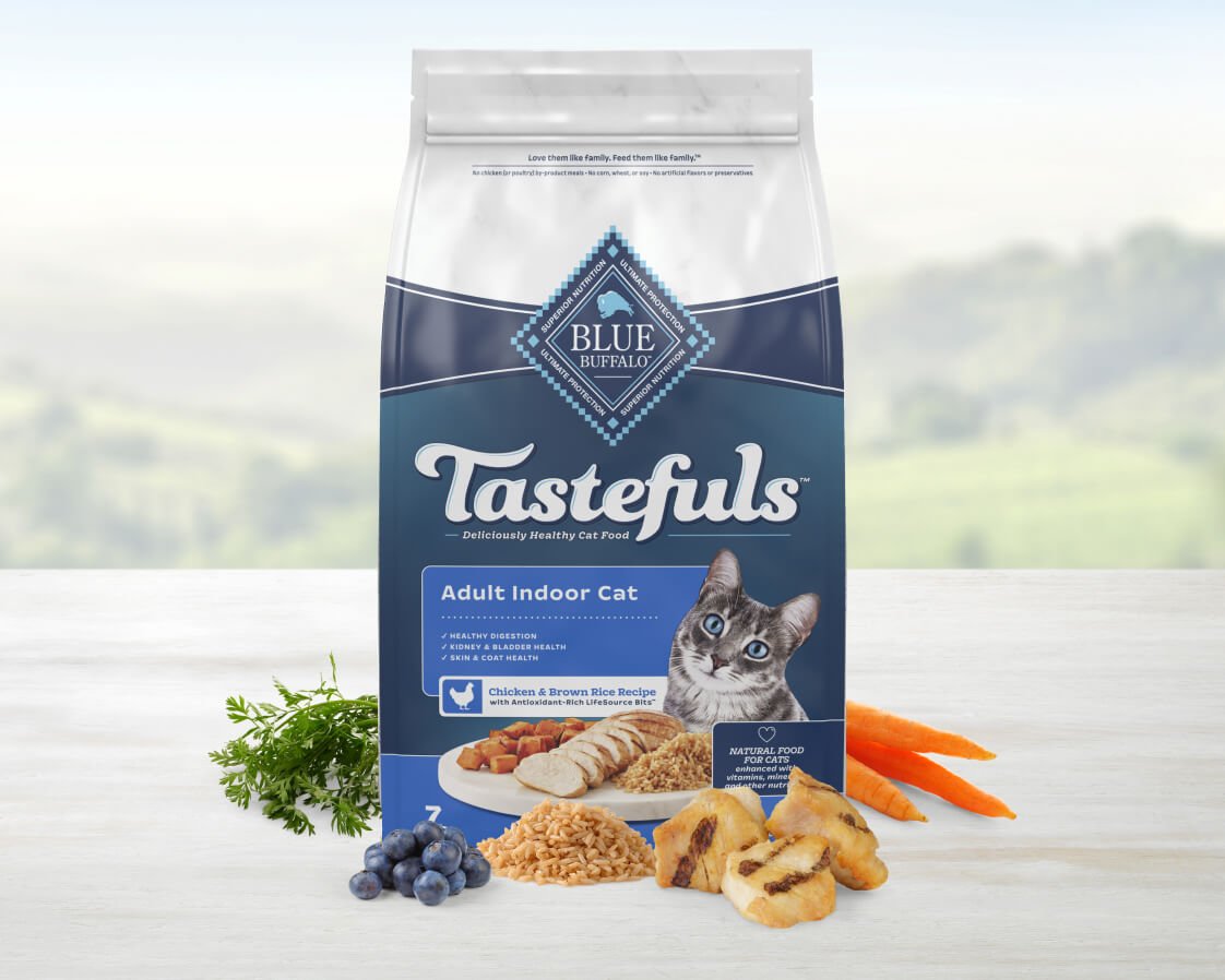 Blue Buffalo Tastefuls adult indoor cat food package on a table, with fresh ingredients like carrots, blueberries, and chicken showcased beside it.