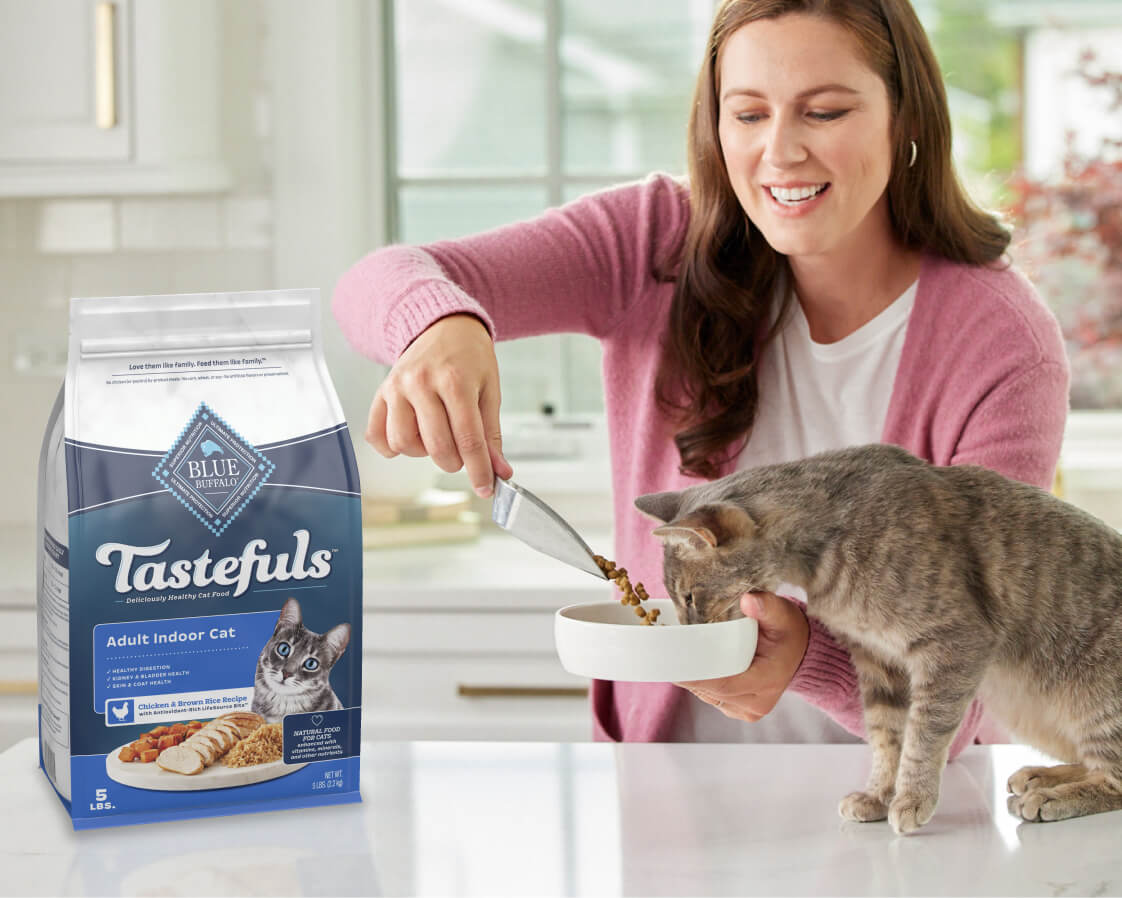 A smiling woman in a pink sweater scoops food from a Blue Buffalo Tastefuls cat food bag into a bowl as a grey tabby cat begins to eat.