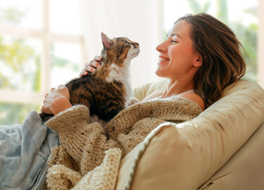 A Pet Parent is smiling as she pets her cat while lounging on the couch under two blankets.