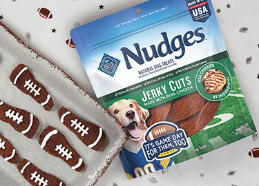 A pack of Nudges Jerky Cuts is next to a tray with Nudges treats with white frosting on them to resemble footballs.