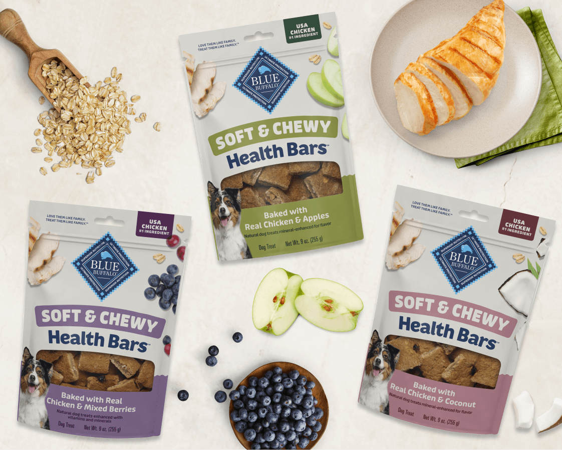 Three bags of BLUE Treats are arranged on a light countertop with ingredients around them. Fresh fruit, a scoop of oats, and a sliced chicken breast on a plate appear in the background.