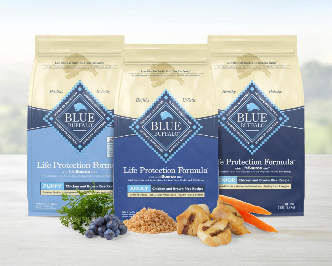 Three Blue Buffalo dog food bags for puppy, adult, and senior stages, with chicken, rice, carrots, and blueberries displayed.