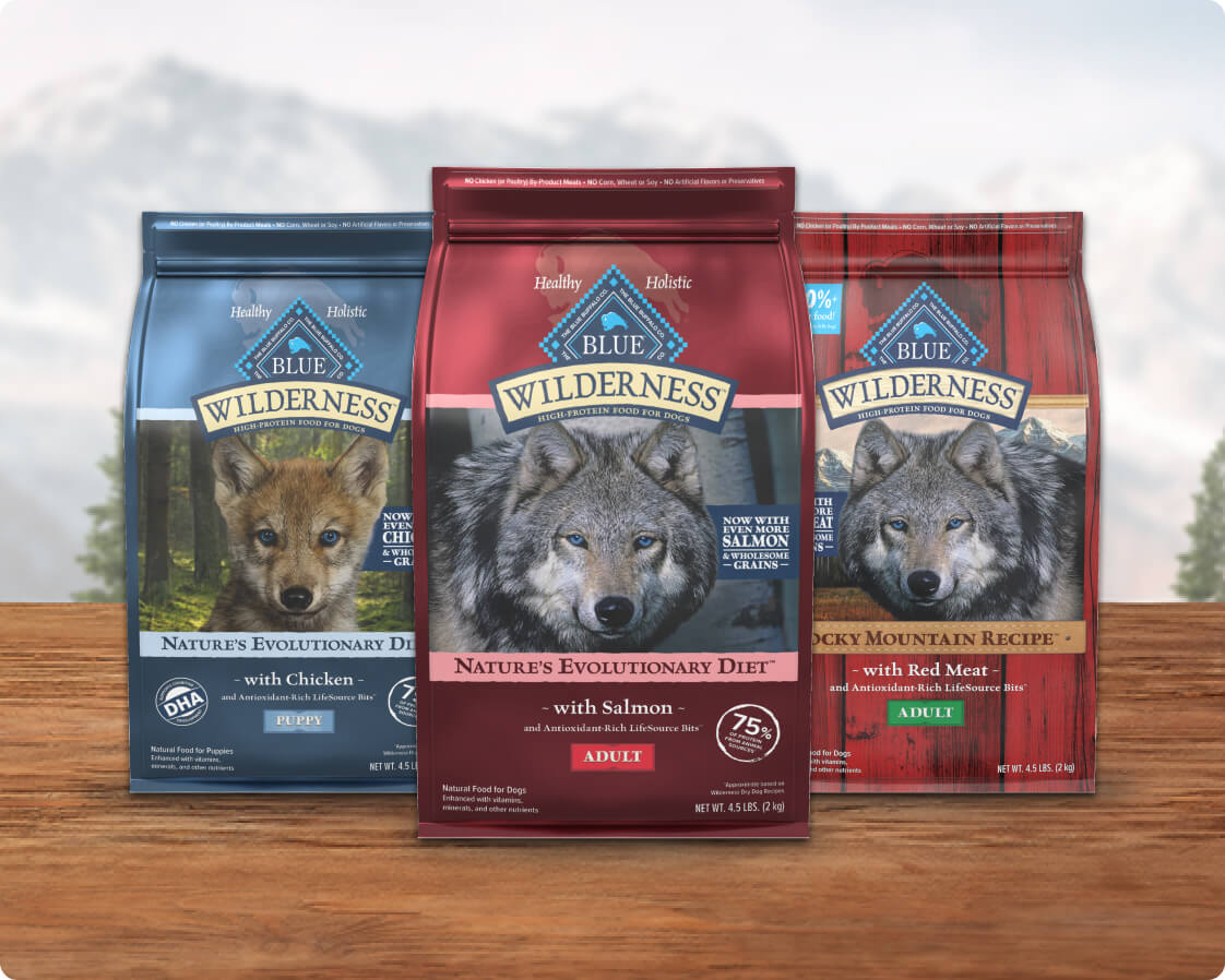 Three bags of Blue Wilderness dog food featuring wolf imagery, labeled with chicken, salmon, and red meat flavors for adults.