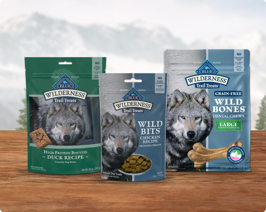 Three packages of Blue Wilderness dog treats are displayed on a wooden surface.