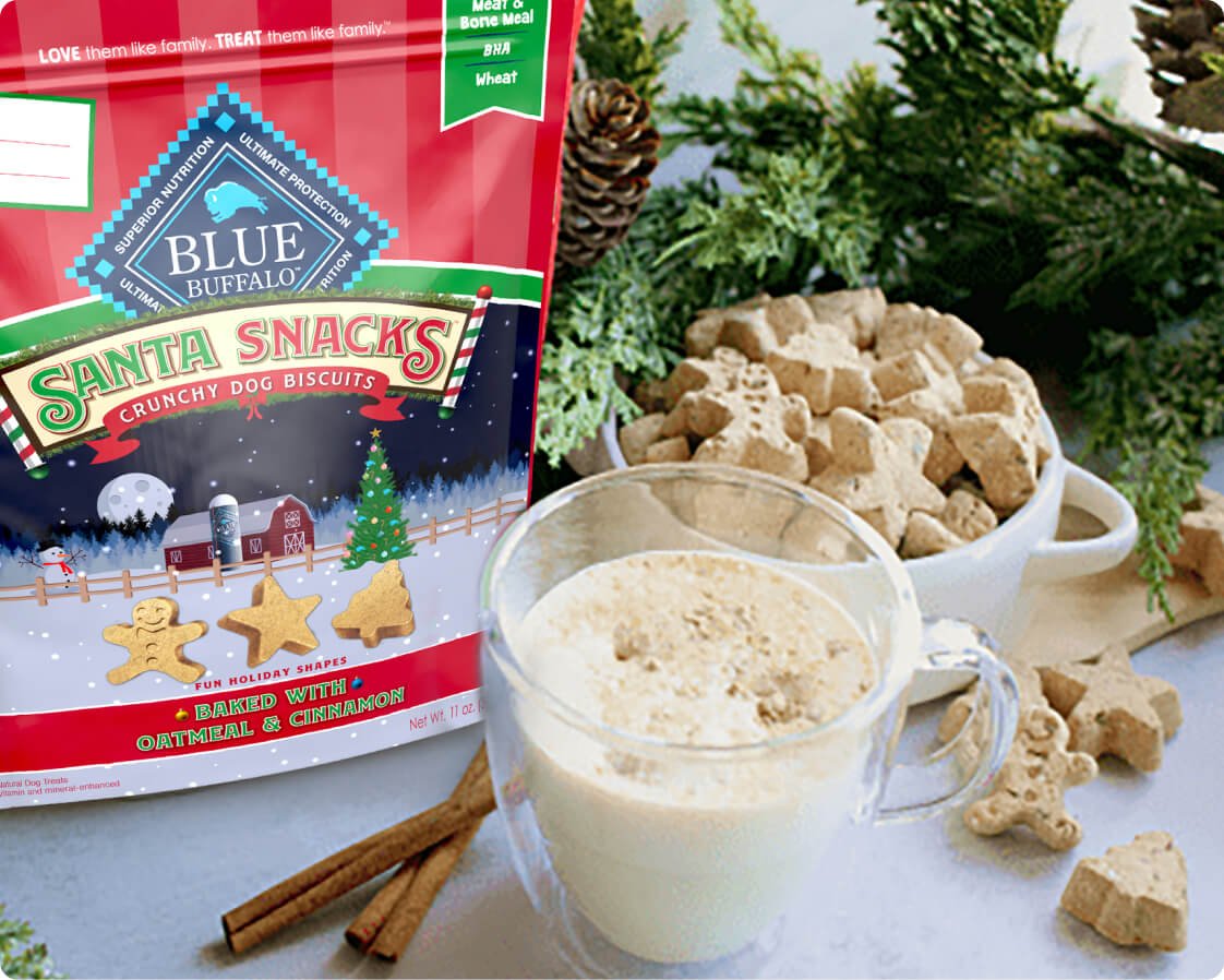 A pack of BLUE Santa Snacks Biscuits is next to a glass mug filled with a dog-friendly gingerbread latte. There are cinnamon sticks and a mug of Santa Snack Biscuits around the glass.