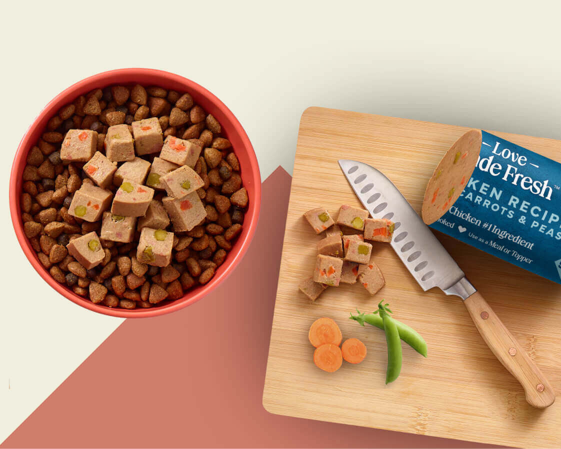 A bowl with dry food & cubes of Love Made Fresh food & a cutting board with cubes of Love Made Fresh, a knife, carrots & peas