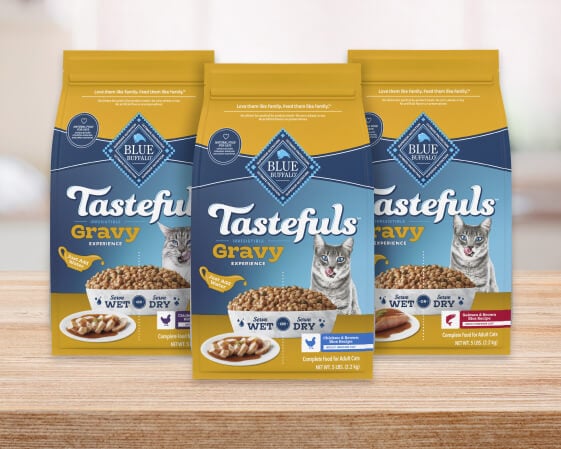 Three bags of BLUE Tastefuls Gravy dry cat food are on a wooden counter.