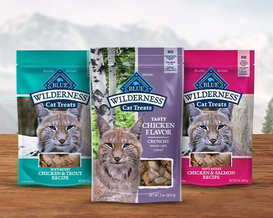 Three packages of Blue Wilderness cat treats in different flavors on a wooden surface.