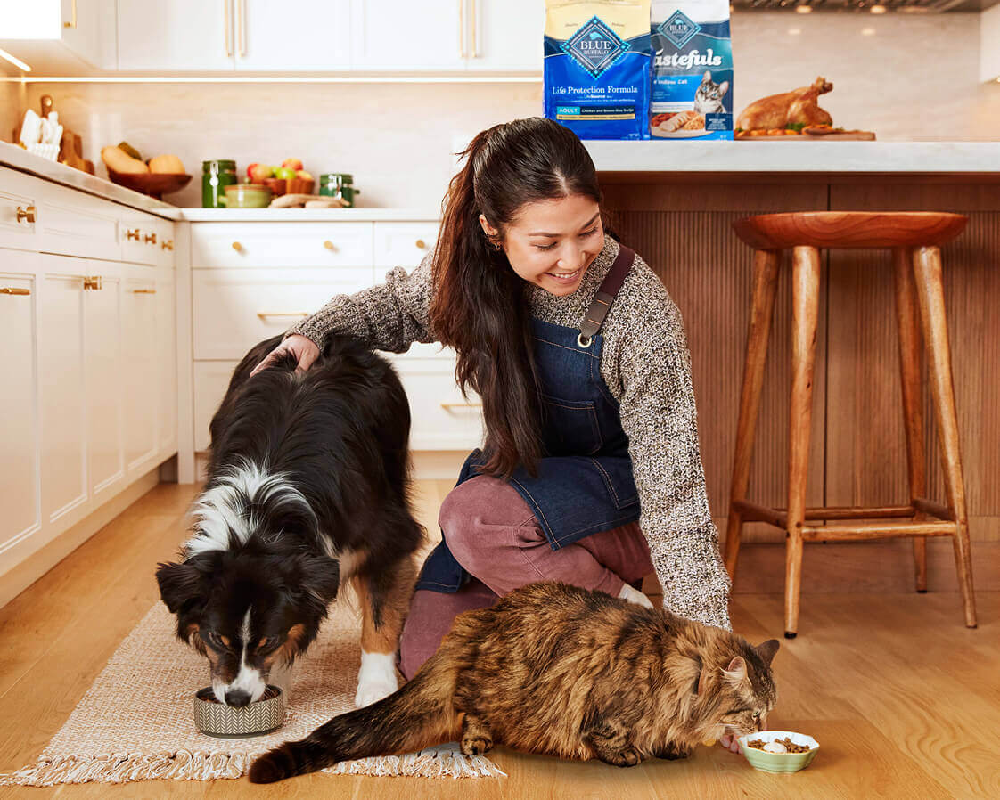 A Pet Parent is smiling and crouching, watching her dog and cat eat in their kitchen.
