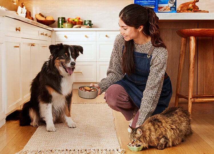 A Pet Parent is smiling and crouching, watching her dog and cat eat in their kitchen.