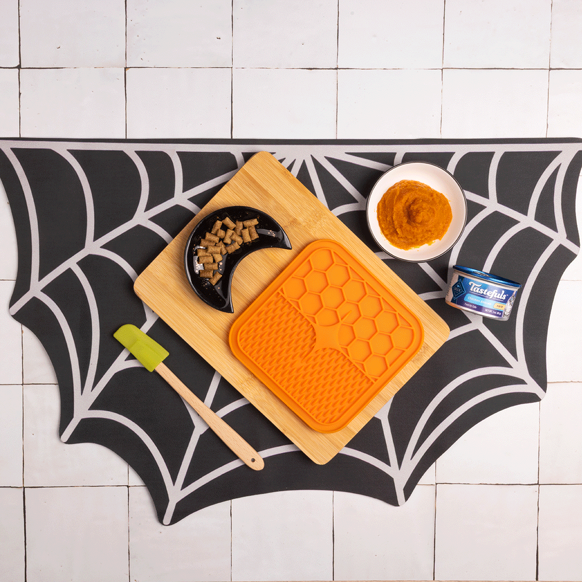 A gif of someone making the recipe on a spider web placemat w/a lick mat, a half moon-shaped bowl, a spatula, & a bowl of Tastefuls cat food