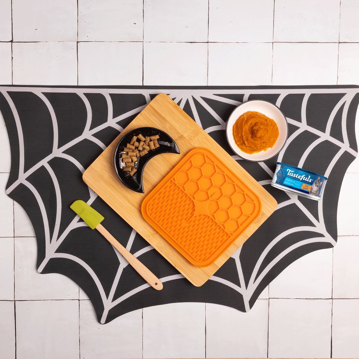 A gif of someone making the recipe on a spider web placemat w/a lick mat, a half moon-shaped bowl, a spatula, & a bowl of Tastefuls cat food