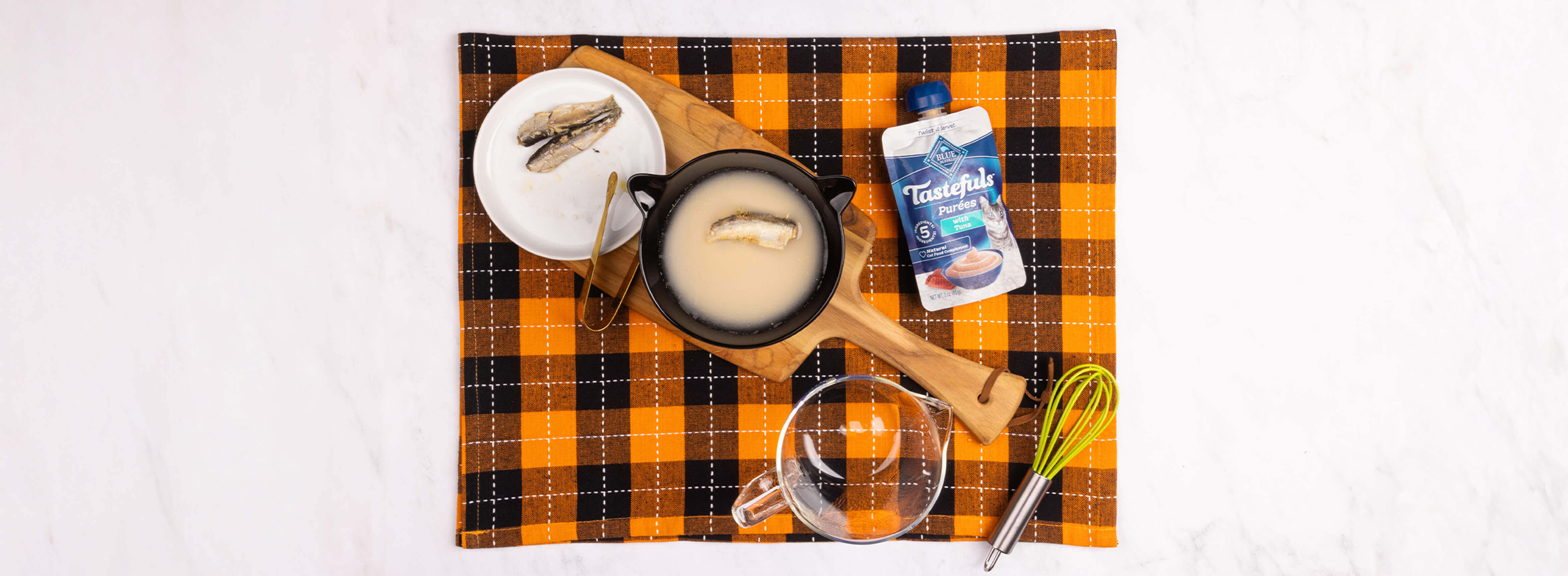 A cat martini using water, a plate w/a sardine, a black bowl, tongs, a Tastefuls purée pouch, a whisk & a glass bowl.