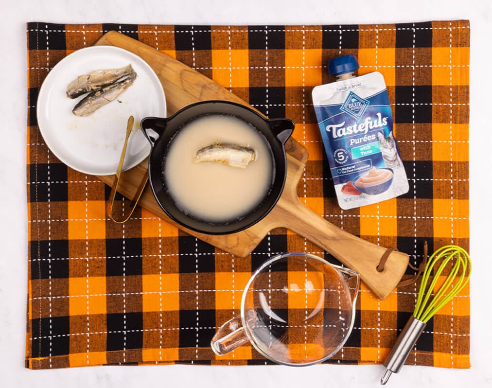 A cat martini using water, a plate w/a sardine, a black bowl, tongs, a Tastefuls purée pouch, a whisk & a glass bowl.