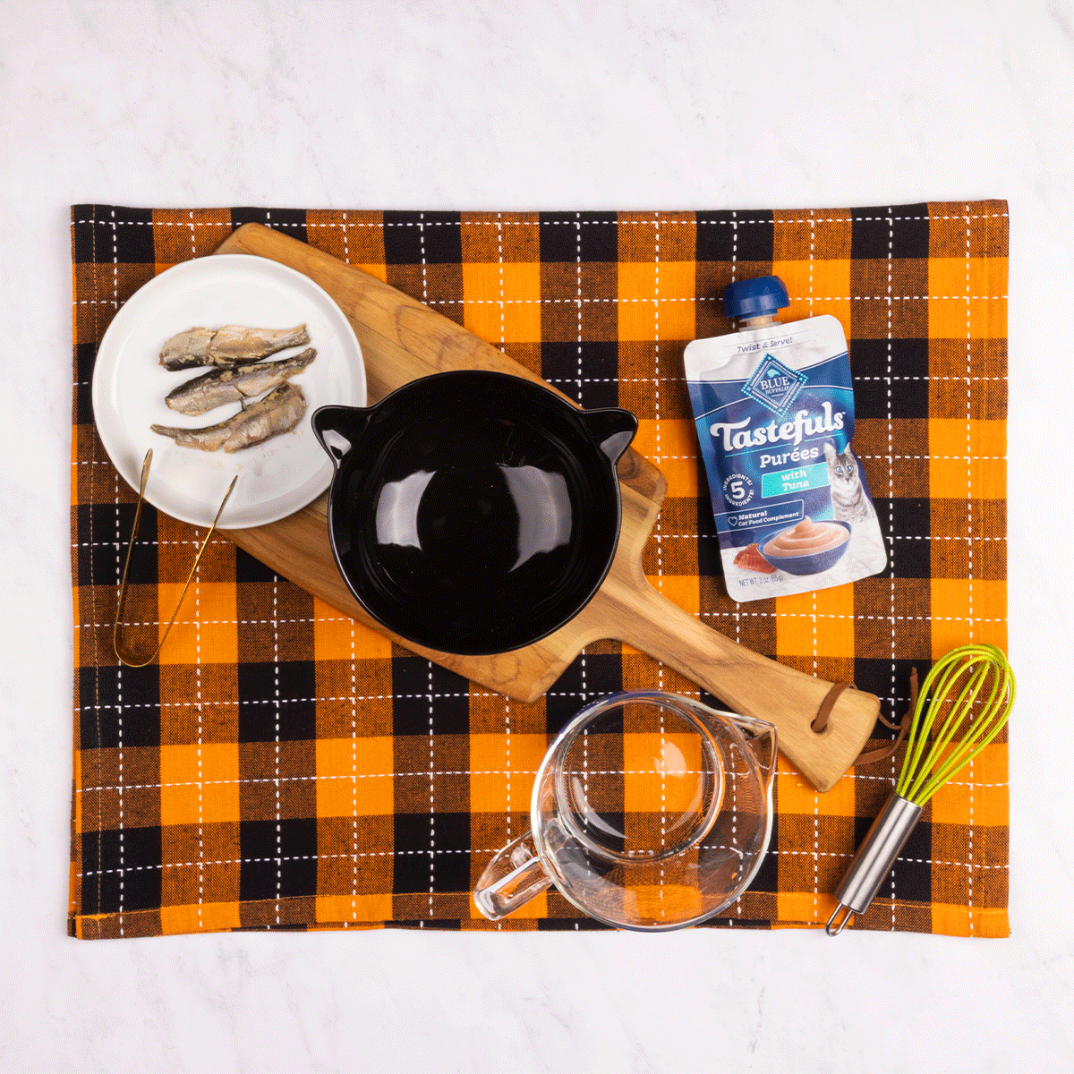 A gif of someone making a cat martini using water, a plate w/a sardine, a black bowl, tongs, a Tastefuls purée pouch, a whisk & a glass bowl.