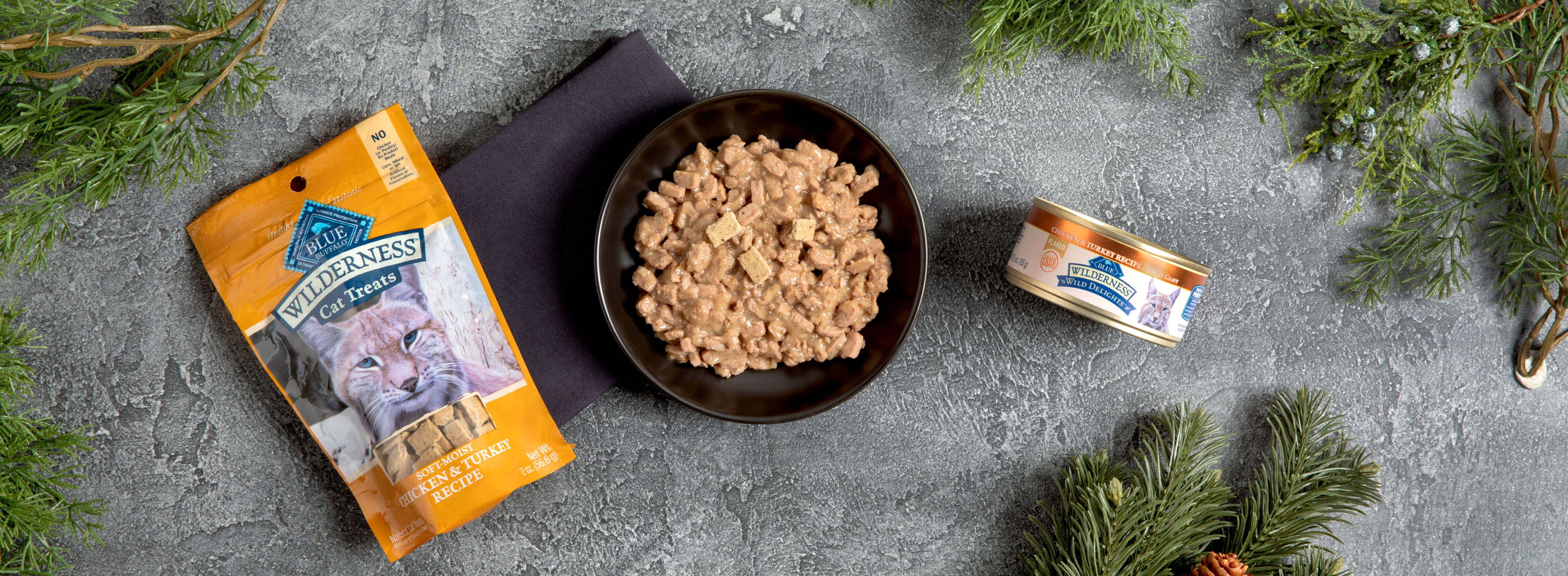 A bowl of wet cat food is flanked by a packet of treats and a can, on a gray surface with pine branches.