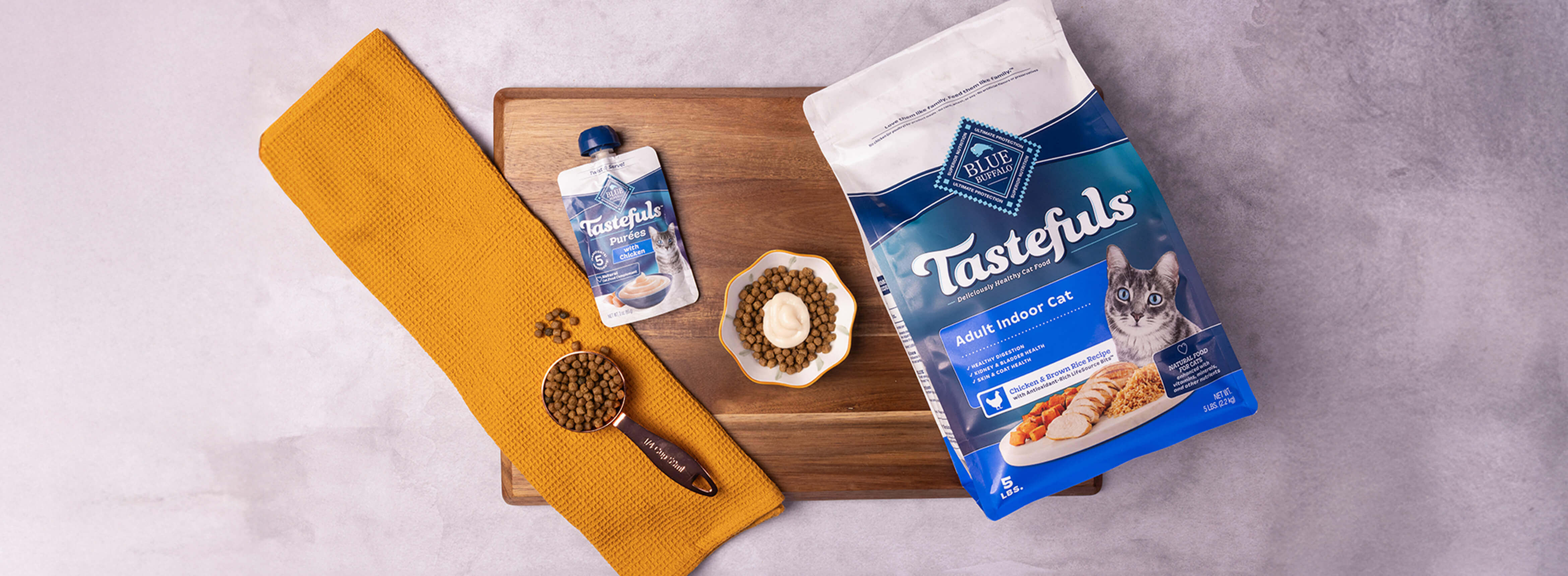 A cat-friendly meal sits in a bowl on a cutting board, surrounded by a pouch of BLUE Tastefuls Purées and a bag of BLUE Tastefuls dry cat food.