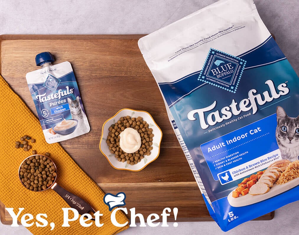 A cat-friendly meal sits in a bowl on a cutting board, surrounded by a pouch of BLUE Tastefuls Purées and a bag of BLUE Tastefuls dry cat food.