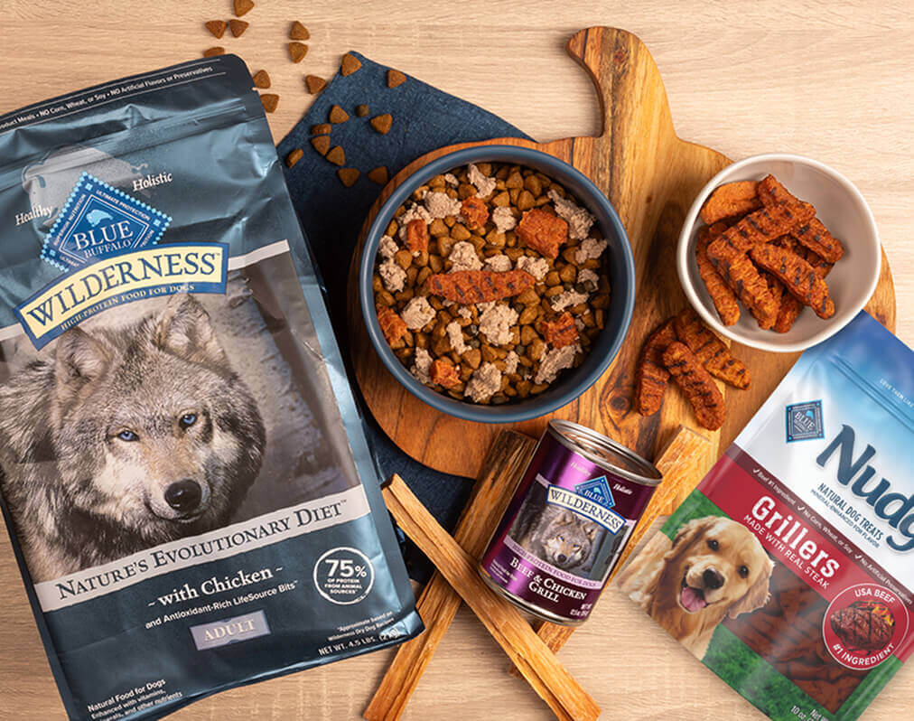 A dog-friendly meal sits in a bowl on a cutting board, surrounded by packages of BLUE Wilderness dry dog food, BLUE Wilderness wet dog food, and BLUE Nudges Grillers.