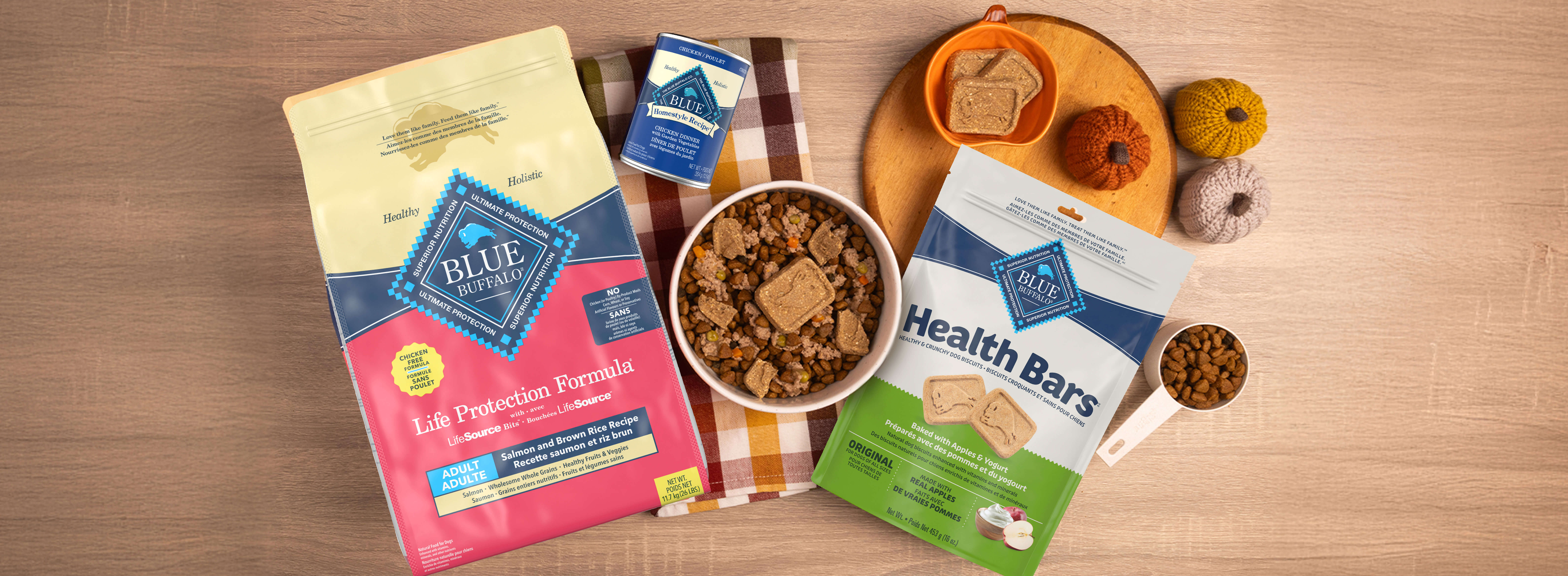 A dog-friendly meal sits in a bowl on a hand towel, surrounded by packages of BLUE Life Protection Formula, BLUE Homestyle Recipe, and BLUE Health Bars, along with autumnal decor.