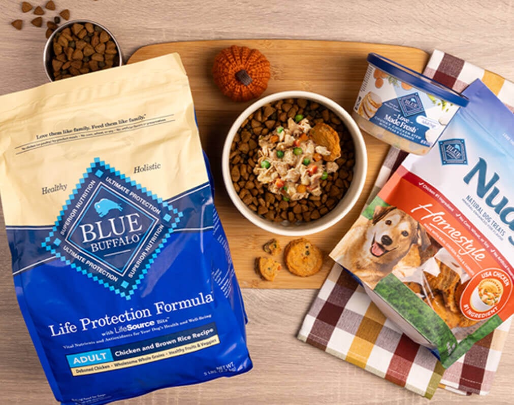 A dog-friendly meal sits in a bowl on a cutting board, surrounded by packages of BLUE Life Protection Formula, BLUE Nudges, and a tub of BLUE Love Made Fresh.