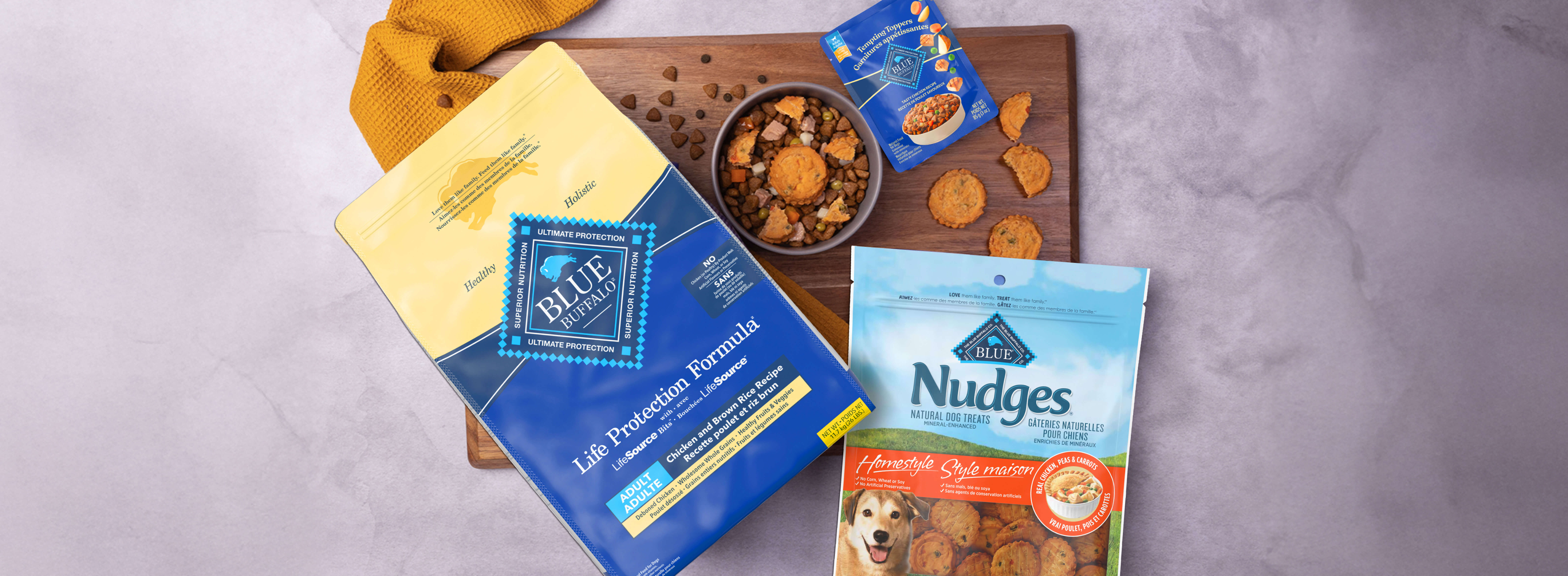 A dog-friendly meal sits in a bowl on a cutting board, surrounded by packages of BLUE Life Protection Formula, BLUE Tempting Toppers, and BLUE Nudges.