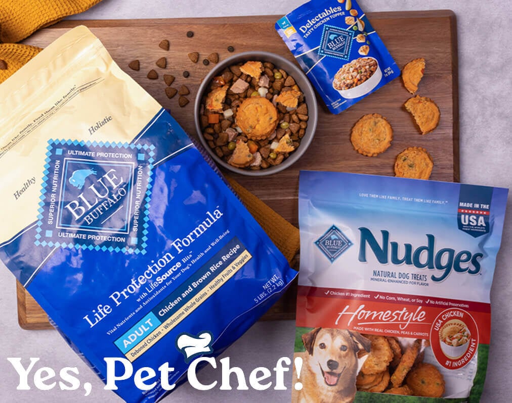 A dog-friendly meal sits in a bowl on a cutting board, surrounded by packages of BLUE Life Protection Formula, BLUE Delectables, and BLUE Nudges.