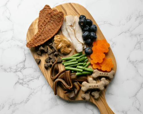 Assorted dog treats and fresh foods such as blueberries, green beans, and sliced chicken, artistically arranged on a wooden board shaped like a dog bone.