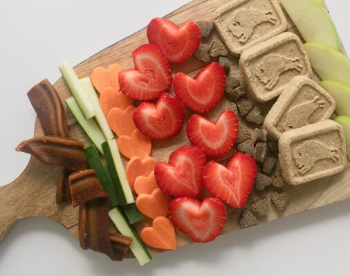 A wooden board with an assortment of dog treats and fruits and vegetables arranged in a visually appealing way.  The treats appear to be Blue Buffalo brand.  The fruits and vegetables include strawberries, apples, carrots, and cucumbers.