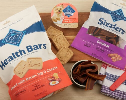 Packages of Blue Buffalo dog treats including Health Bars and Sizzlers, alongside a bowl of treats and a dog toy, arranged on a wooden surface.