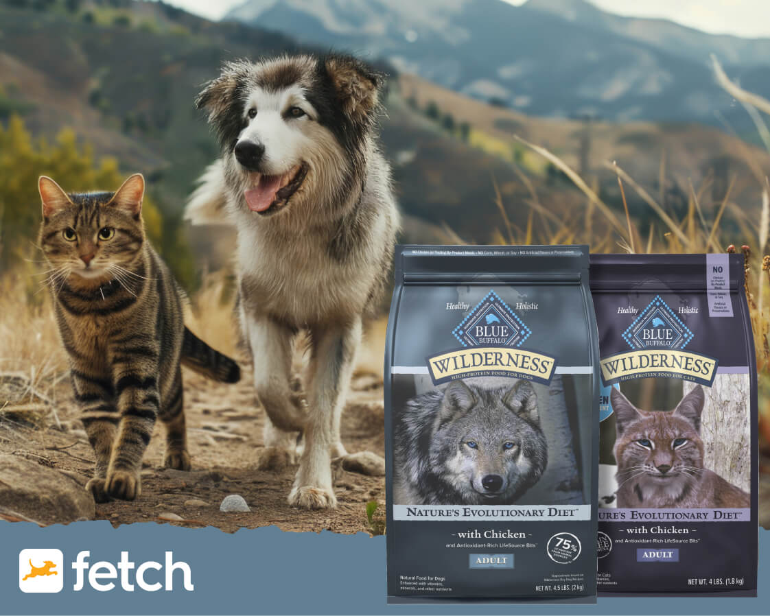 A cat and dog outdoors with two Blue Wilderness pet food bags, mountains rising in the background.