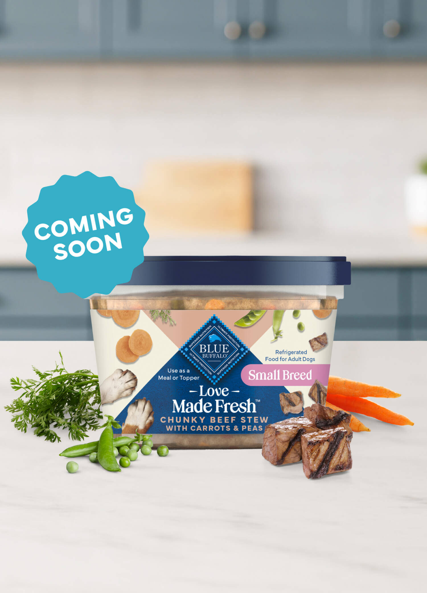 A tub of Love Made Fresh beef stew is surrounded by parsley, chunks of seared beef, carrots, and peas on a countertop.