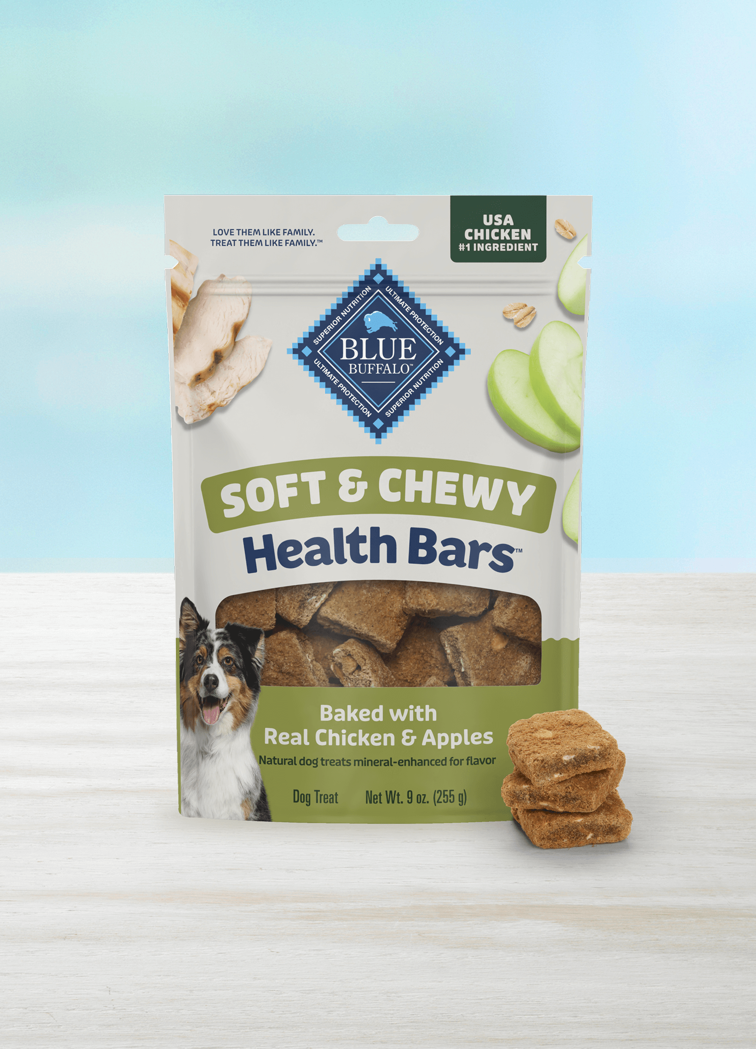 A bag of BLUE dog treats sits on a light surface against a blue-and-white background. The package shows crispy, sliced apples, pieces of juicy tender chicken, and a happy dog. A small stack of treats sits beside the treat bag.