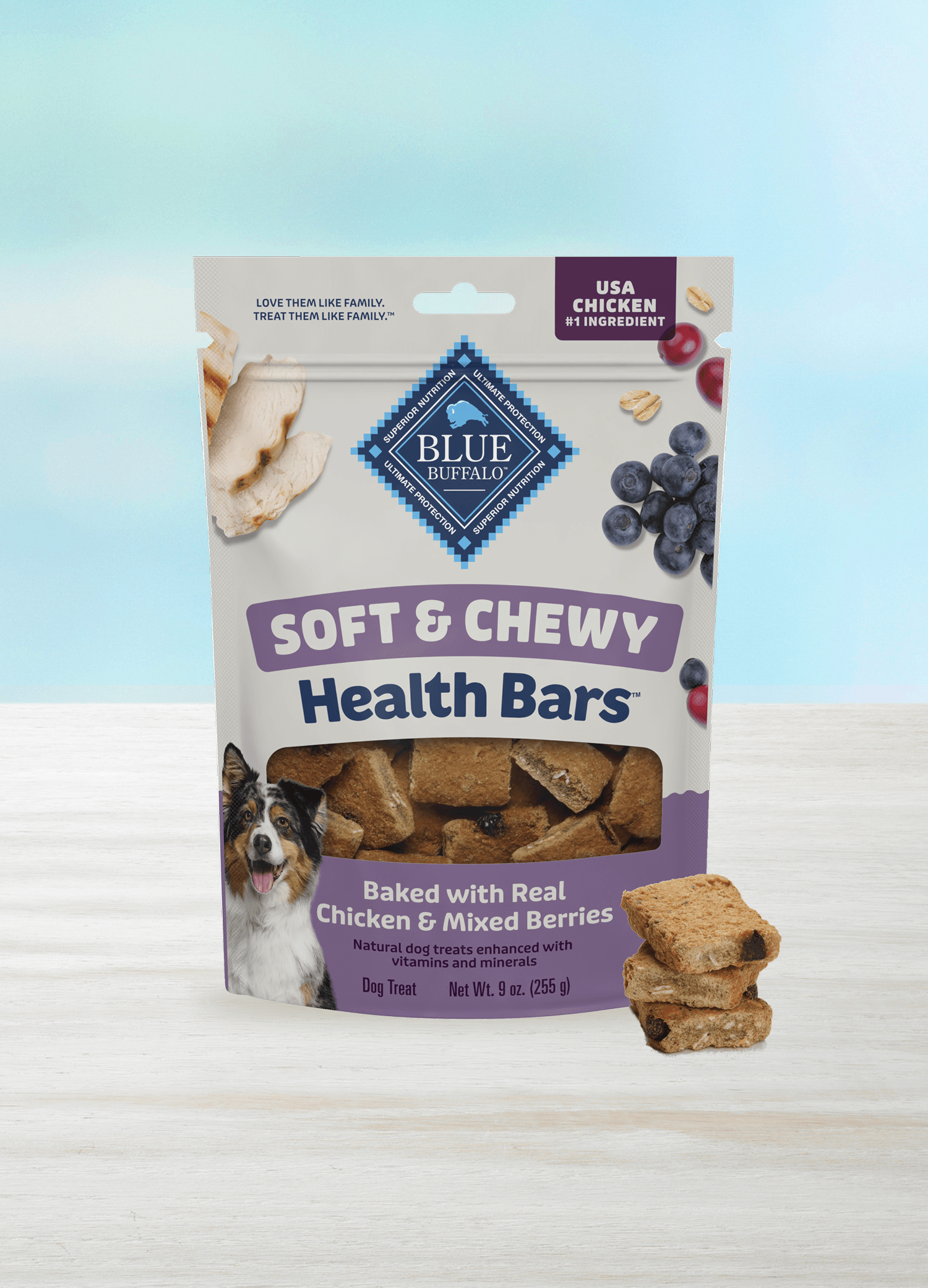 A bag of BLUE dog treats sits on a light surface against a blue-and-white background. The package shows juicy berries, pieces of tender chicken, and a happy dog. A small stack of treats sits beside the treat bag.